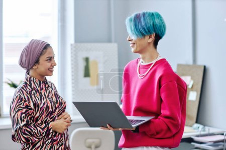 Photo for Side view portrait of two gen Z young people chatting while standing in office and holding laptop - Royalty Free Image