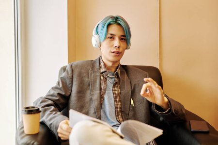 Téléchargez les photos : Portrait de jeune homme asiatique avec des cheveux colorés portant des écouteurs et regardant la caméra tout en se relaxant au bureau - en image libre de droit