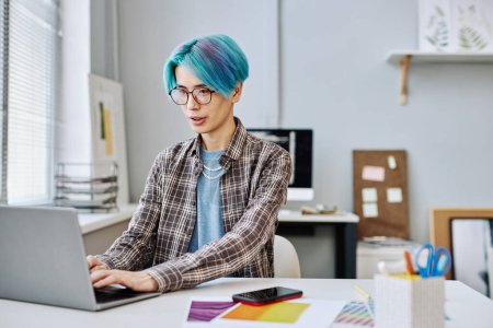 Téléchargez les photos : Portrait de jeune homme créatif aux cheveux bleus utilisant un ordinateur portable au bureau, espace de copie - en image libre de droit