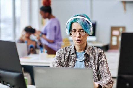 Téléchargez les photos : Portrait de jeune homme asiatique aux cheveux bleus utilisant l'ordinateur au bureau et portant des écouteurs - en image libre de droit