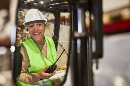 Téléchargez les photos : Portrait d'une jeune femme souriante portant un casque de sécurité lors de l'utilisation d'un chariot élévateur dans un entrepôt, espace de copie - en image libre de droit