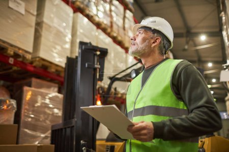 Photo for Side view portrait of bearded worker wearing hardhat and holding clipboard while managing warehouse, copy space - Royalty Free Image
