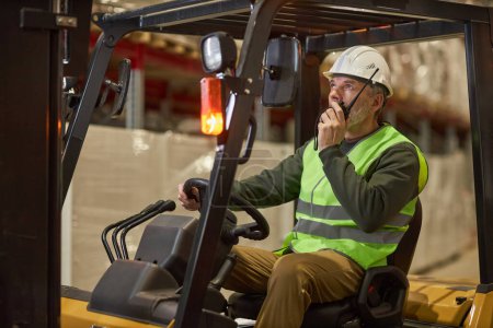 Téléchargez les photos : Portrait d'un travailleur masculin conduisant un chariot élévateur dans un entrepôt et parlant à une radio portable - en image libre de droit