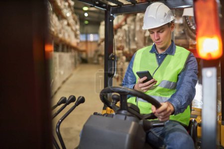 Téléchargez les photos : Portrait de jeune travailleur conduisant un chariot élévateur dans un entrepôt et utilisant un smartphone - en image libre de droit