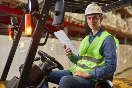 Photo for Side view portrait of young male worker driving forklift truck in warehouse and smiling at camera - Royalty Free Image