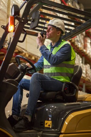 Photo for Vertical portrait of young male worker driving forklift truck in warehouse and speaking to portable radio - Royalty Free Image