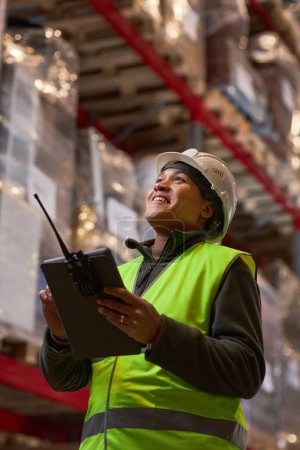 Photo for Vertical portrait of black woman wearing hardhat in storage warehouse and smiling happily - Royalty Free Image