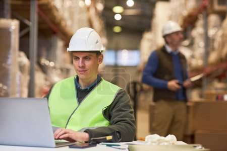 Téléchargez les photos : Portrait d'un travailleur souriant utilisant un ordinateur portable dans un entrepôt tout en gérant un système de distribution - en image libre de droit