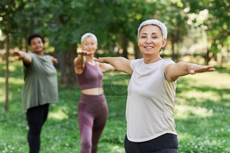 Téléchargez les photos : Portrait taille haute de femme âgée souriante appréciant l'entraînement sportif à l'extérieur et regardant la caméra tout en faisant de l'exercice d'étirement, copier l'espace - en image libre de droit