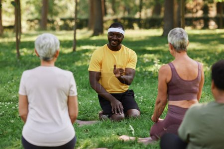 Téléchargez les photos : Portrait complet d'un entraîneur de yoga souriant parlant à un groupe de femmes âgées en séance d'entraînement en plein air - en image libre de droit