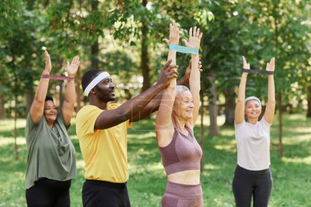 Téléchargez les photos : Vue latérale de l'instructeur de sport masculin aidant les femmes âgées à s'entraîner avec des élastiques dans le parc - en image libre de droit