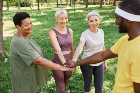 Téléchargez les photos : Groupe de femmes âgées actives empilant les mains avec un entraîneur après avoir apprécié l'entraînement en plein air dans le parc - en image libre de droit