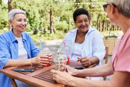 Foto de Diverso grupo de mujeres mayores disfrutando de bebidas en la cafetería al aire libre y sonriendo felizmente - Imagen libre de derechos