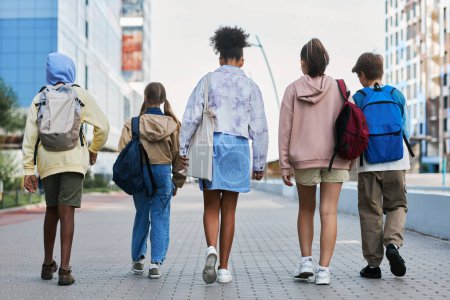 Téléchargez les photos : Vue arrière de jeunes élèves interraciaux de l'école secondaire avec des sacs à dos marchant dans la rue tout en revenant à la maison après les cours - en image libre de droit