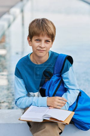 Photo for Cute secondary school learner with open book looking at camera while sitting in urban environment and preparing for lesson of literature - Royalty Free Image