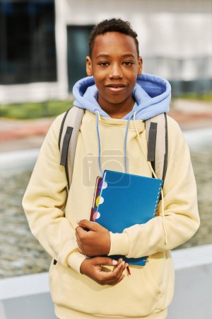 Foto de Sonriente colegial afroamericano con copybooks y mochila de pie frente a la cámara en el entorno urbano - Imagen libre de derechos