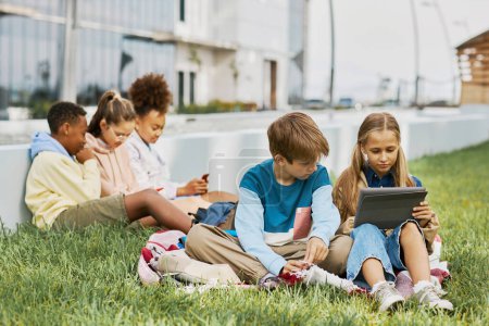 Téléchargez les photos : Deux écoliers adolescents regardant l'écran de la tablette tenue par la fille tout en étant assis sur l'herbe verte et en regardant la vidéo en ligne contre leurs amis - en image libre de droit