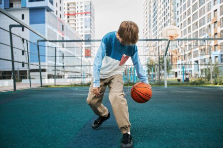 Téléchargez les photos : Apprenante active du secondaire en pantalon et pull-over jouant au basket sur terrain de sport après les cours ou à loisir contre les maisons - en image libre de droit