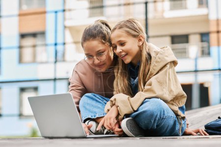Téléchargez les photos : Deux écolières heureuses en tenue décontractée regardant un écran d'ordinateur portable tout en regardant un film en ligne ou en communiquant en chat vidéo à l'extérieur - en image libre de droit