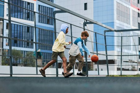 Foto de Dos chicos multiculturales en ropa deportiva corriendo tras la pelota y tratando de atraparla mientras juegan baloncesto en el ocio o después de la escuela al aire libre - Imagen libre de derechos