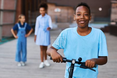 Téléchargez les photos : Un garçon afro-américain souriant en t-shirt bleu clair chevauchant un scooter électrique et regardant la caméra contre deux filles interculturelles - en image libre de droit