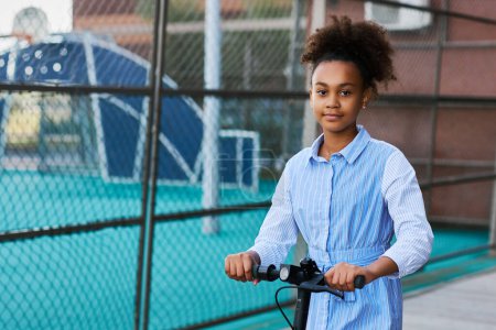Téléchargez les photos : Jolie adolescente afro-américaine avec scooter électrique regardant la caméra tout en restant près de l'aire de jeux avec des installations sportives - en image libre de droit