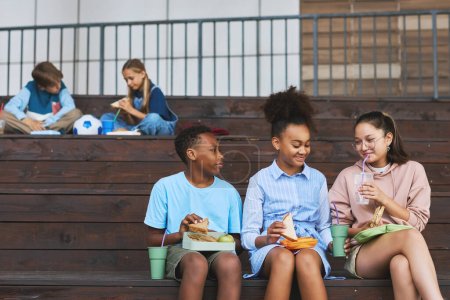 Téléchargez les photos : Groupe de trois amis d'école multiculturels heureux ayant sandwichs et soda pour le déjeuner tout en étant assis en rangée sur un escalier en bois - en image libre de droit