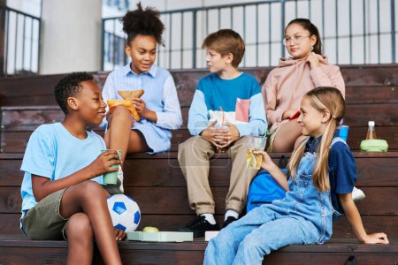 Foto de Jóvenes amigos de la escuela intercultural con refrescos mirándose mientras están sentados en una escalera de madera y charlando contra otros niños - Imagen libre de derechos