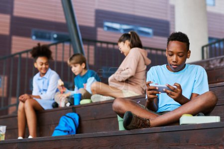 Téléchargez les photos : Adolescent afro-américain écolier en casual utilisant un smartphone tout en étant assis sur un escalier en bois contre un groupe d'amis d'école - en image libre de droit