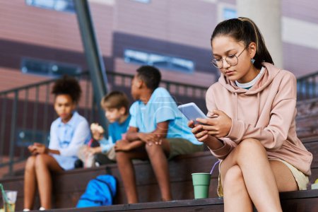 Photo for Youthful pretty schoolgirl in casualwear and eyeglasses texting in smartphone in front of camera with her friends on background - Royalty Free Image