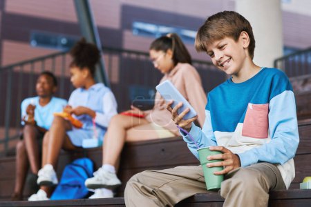 Téléchargez les photos : Heureux écolier adolescent avec verre de soude en regardant l'écran du smartphone tout en regardant la vidéo en ligne contre ses amis - en image libre de droit