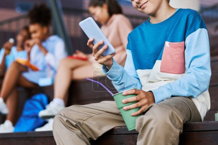 Foto de Primer plano del adolescente en ropa casual mensajes de texto en el teléfono móvil y tener refrescos mientras está sentado frente a la cámara contra sus amigos - Imagen libre de derechos