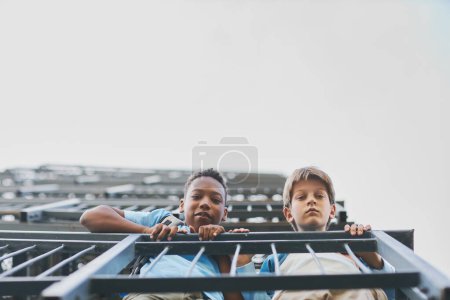 Zwei niedliche interkulturelle Jugendliche gucken vom Balkon eines Wolkenkratzers, während sie sich an Metallgeländern festhalten und in die Kamera schauen