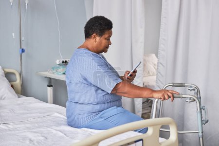 Photo for Side view portrait of black senior woman using phone while sitting alone in hospital room, copy space - Royalty Free Image