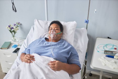 Foto de Retrato de alto ángulo de mujer mayor negra acostada en la cama en la habitación del hospital con máscara de soporte de oxígeno - Imagen libre de derechos
