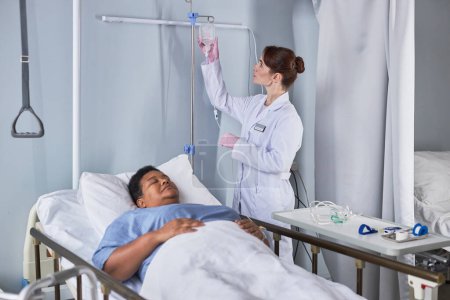 Foto de Retrato de la joven enfermera que establece la terapia IV para la mujer mayor en la cama de hospital - Imagen libre de derechos