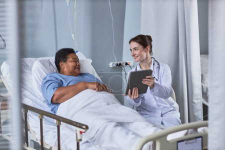 Foto de Retrato de una joven enfermera sonriente mostrando una tableta digital a una mujer mayor en la habitación del hospital - Imagen libre de derechos