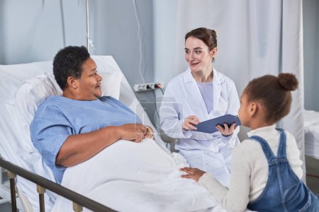 Téléchargez les photos : Portrait d'une jeune infirmière souriante parlant à une femme âgée dans une chambre d'hôpital - en image libre de droit