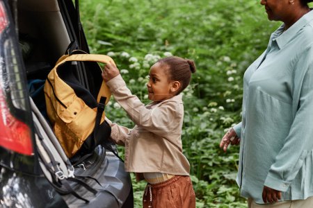 Téléchargez les photos : Vue latérale portrait de la petite fille noire mettant sac à dos dans le coffre de la voiture tout en voyageant avec maman - en image libre de droit