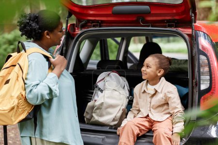 Téléchargez les photos : Portrait de mignonne fille noire assise dans le coffre de la voiture et profiter de voyager avec la mère - en image libre de droit
