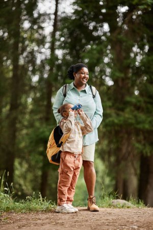 Photo for Vertical portrait of black little girl enjoying hiking in nature trail and looking in binoculars with mom - Royalty Free Image
