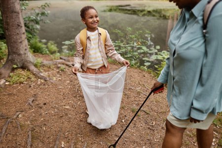 Téléchargez les photos : Portrait grand angle de fille noire souriante tenant sac poubelle à l'extérieur et aidant à nettoyer la nature avec maman - en image libre de droit