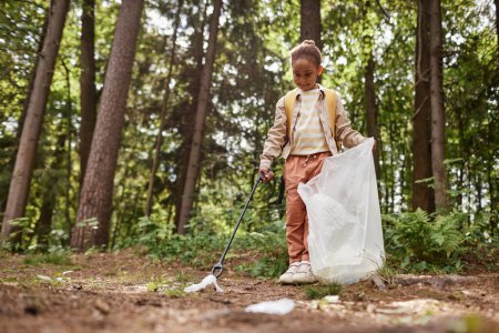 Téléchargez les photos : Portrait complet d'une petite fille souriante ramassant des ordures dans le sentier de la nature, espace de copie - en image libre de droit