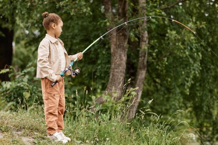 Photo for Full length side view portrait of black little girl fishing by river in nature with fishing rod bending, copy space - Royalty Free Image
