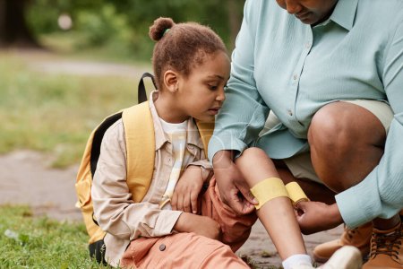 Téléchargez les photos : Portrait de petite fille noire blessée lors d'une randonnée dans la nature avec sa mère mettant un bandage sur le genou à l'extérieur - en image libre de droit