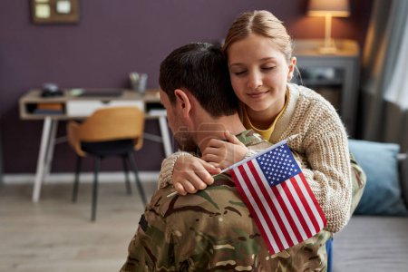 Téléchargez les photos : Portrait d'une adolescente embrassant un père militaire et tenant un drapeau américain à la maison, espace de copie - en image libre de droit