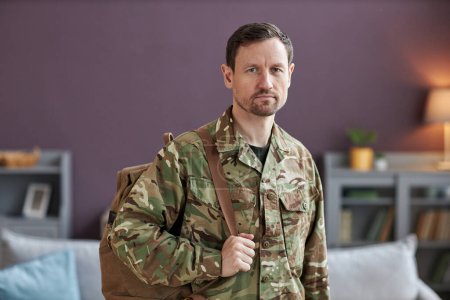 Foto de Cintura hacia arriba retrato militar hombre sosteniendo la mochila y mirando a la cámara en el entorno del hogar - Imagen libre de derechos