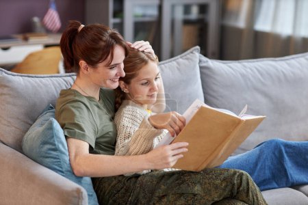 Téléchargez les photos : Vue de côté portrait de femme militaire avec fille à la maison lecture livre ensemble et détente sur canapé - en image libre de droit