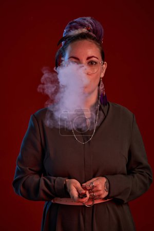 Foto de Mujer joven con cigarrillo electrónico en las manos soplando nube de humo mientras está de pie frente a la cámara contra el fondo rojo oscuro - Imagen libre de derechos