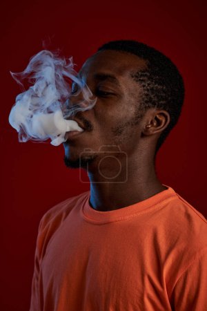 Foto de Joven hombre afroamericano se eleva delante de la cámara mientras fuma cigarrillo electrónico o narguile y sopla nube de humo grueso - Imagen libre de derechos